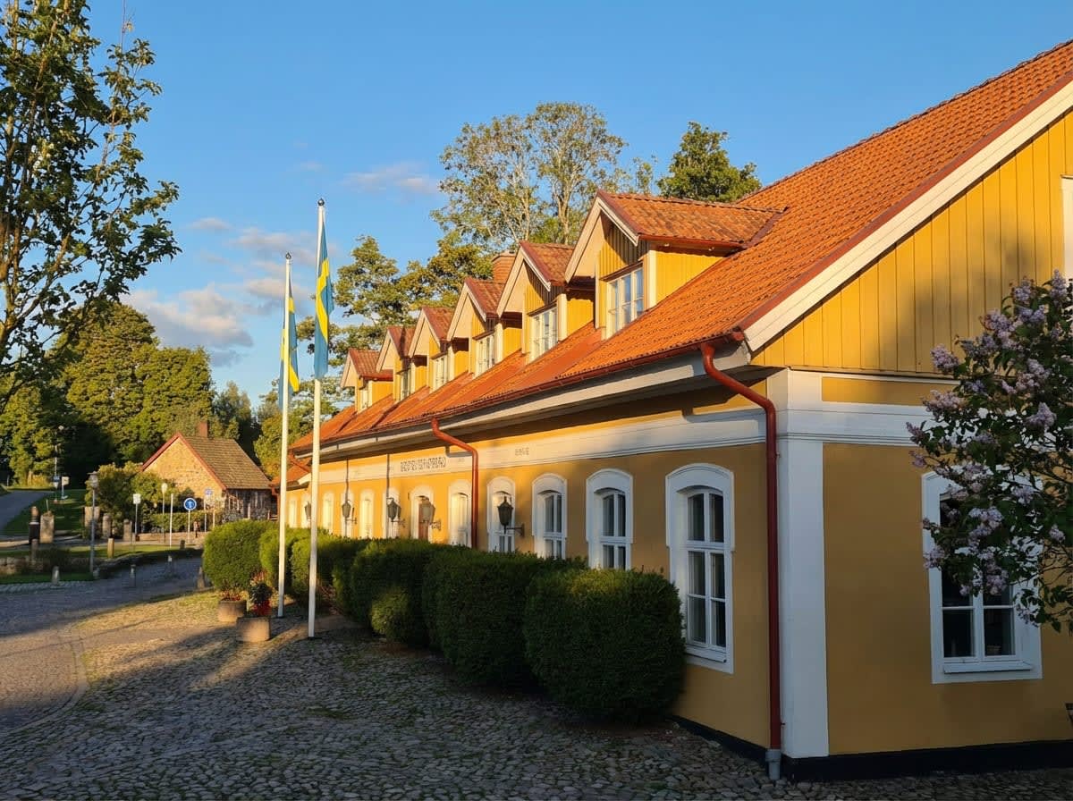 En gul byggnad med rött tak och vita detaljer står vid en kullerstensbelagd yta under en klarblå himmel. Framför byggnaden finns en rad med putsade buskar och två flaggstänger med svenska flaggor. I bakgrunden syns träd.