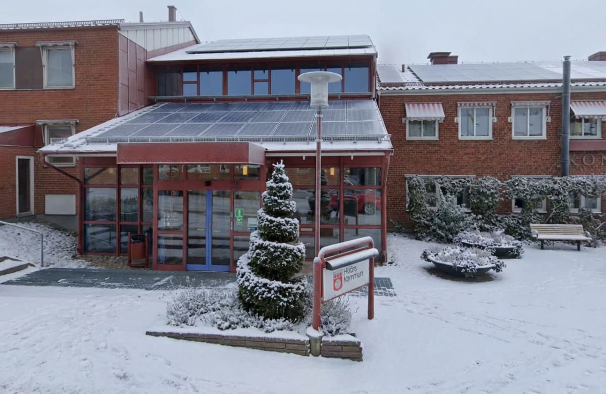 En röd tegelbyggnad delvis täckt av snö på marken och taket. Byggnaden har fönster, solpaneler på taket, en entrédörr av glas och en skylt med texten "Hjörs Kommun". Framför byggnaden syns ett snötäckt träd, en gatlykta och en bänk.