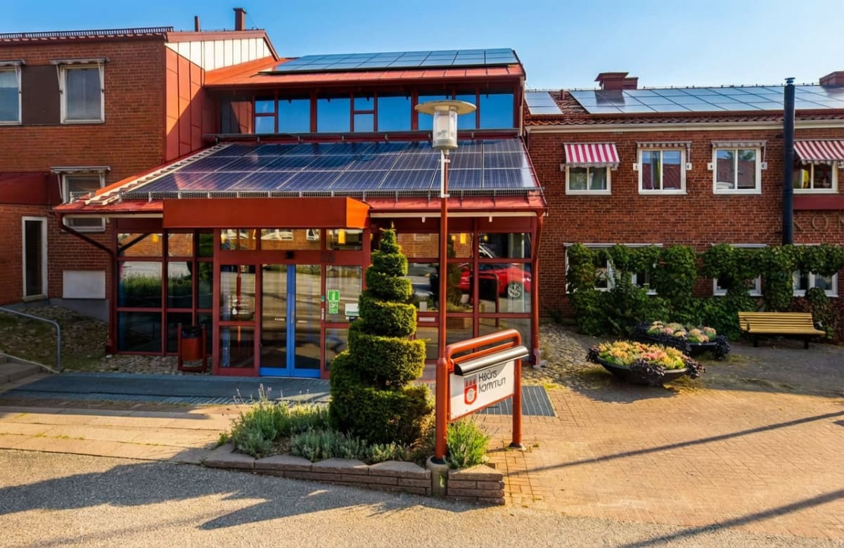 En röd tegelbyggnad med stora glaspartier och solpaneler på taket. Framför byggnaden finns gångplattor, planteringar, en formklippt buske, en gatlampa och en skylt med texten "Hörby kommun". Solen skiner och himlen är klarblå.