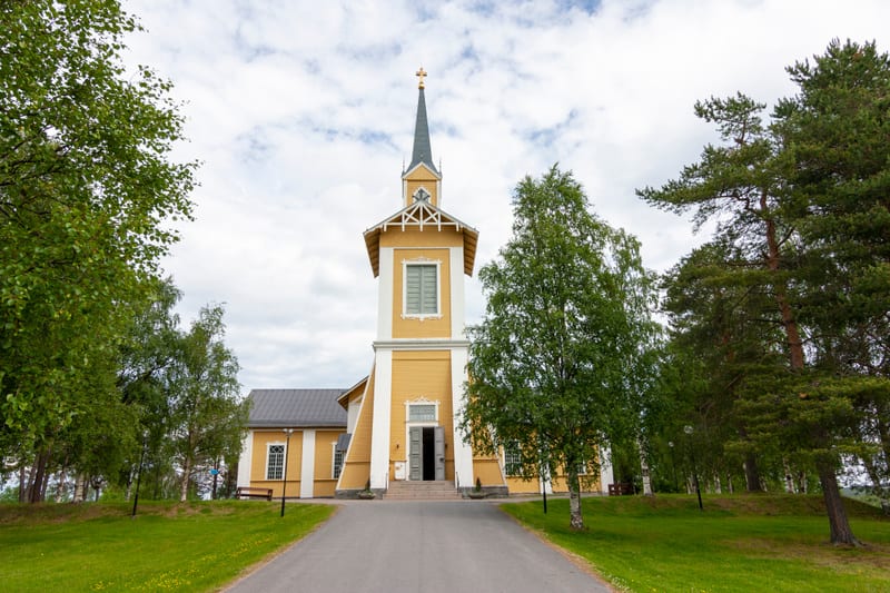 En gul kyrkobyggnad med vitt torn och svart spira, omgiven av gröna träd och gräs. En asfalterad gång leder fram till kyrkans dörrar. Himmelen är ljus med moln. Två bänkar syns vid sidan av byggnaden.