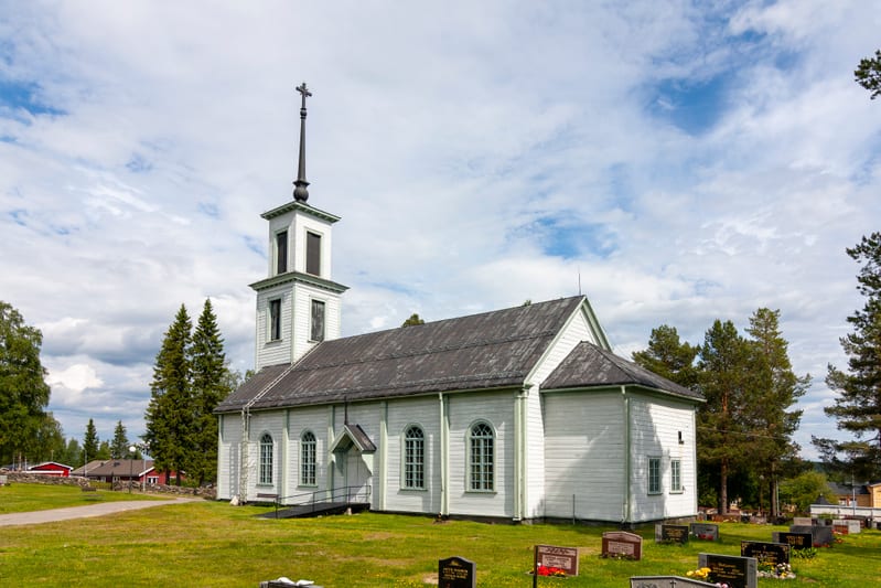 En vit träkyrka med ett smalt torn och en spira med ett kors på toppen, belägen bredvid en kyrkogård med gräsmattor och gravstenar. Bakgrunden visar en delvis molnig blå himmel och barrträd, samt röda byggnader i fjärran. Kyrkan har flera fönster län