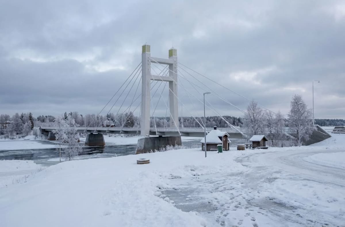 En vinterbild visar en vajerbro över ett delvis isbelagt vattendrag. Omgivningen är täckt av snö, och frostnupna träd kantar floden. På förgrunden, till höger, finns snötäckta utomhustoaletter och en bänk, samt snötäckta ytor med fotspår och spår av