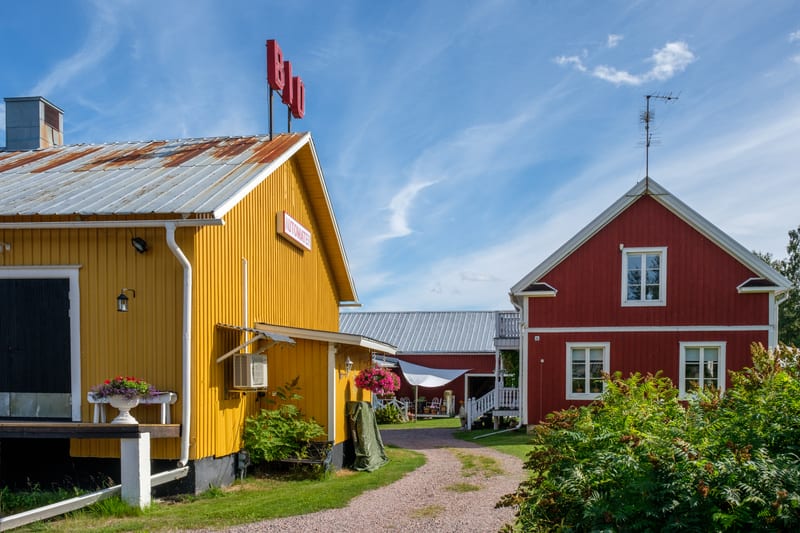 En bild som visar en grusgång mellan två byggnader med färgade fasader och plåttak. Den vänstra byggnaden är gul med en röd skylt som det står "Bio" på taket och "Bioautomaten" på fasaden, medan den högra är röd. Hela scenen är utomhus under en blå h