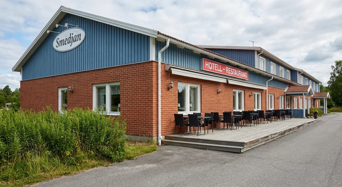 En röd tegelbyggnad med blå träpaneler pryds av skylten "Smedjan". På framsidan finns en uteservering med svarta stolar och bord på en träterrass. En skylt med texten "HOTELL - RESTAURANG" syns ovanför uteserveringen. Bilder i bakgrunden är träd och