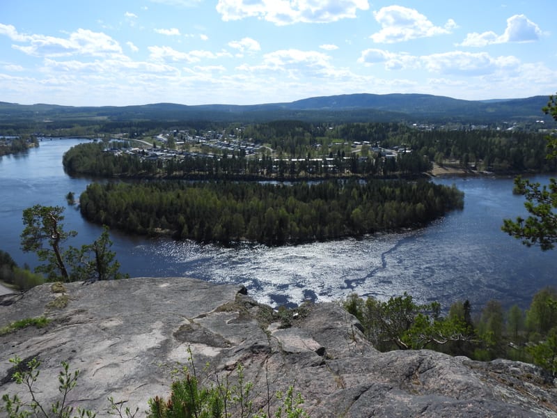 En vidvinkelbild tagen från hög höjd som visar en flod med en stor skogstäckt ö i mitten. På flodens bortre strand syns en bebyggelse omgiven av skog. En del av klippan där bilden är tagen från syns i förgrunden.