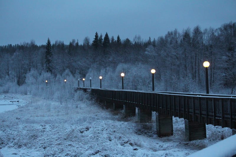 En snötäckt landskapsbild tagen vid skymning eller gryning, med en träbro i förgrunden som sträcker sig diagonalt från höger i bild mot vänster, försedd med flera tända, runda gatlyktor. I bakgrunden syns en skog med snöklädda träd.