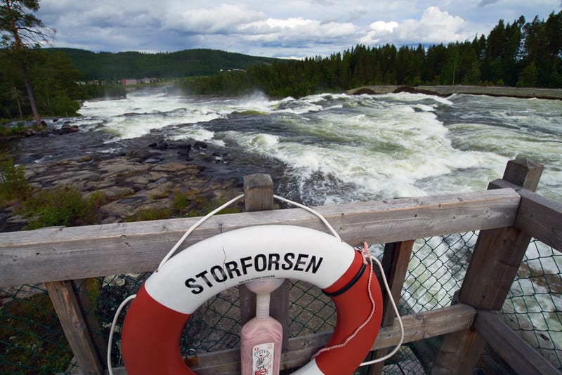 En livboj med texten "STORFORSEN" hänger på ett trästaket i förgrunden. I bakgrunden syns ett skummande vattendrag omgivet av steniga stränder och grön skog. Himmel med moln.