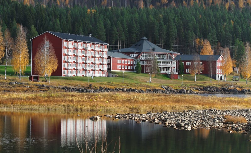 En bild som visar byggnader med röda fasader och vita detaljer vid en flod eller sjö med steniga stränder. Bakom byggnaderna breder en skog ut sig. Träd vid vattnet har gulnat, vilket tyder på höst. Byggnaderna reflekteras i vattnet.
