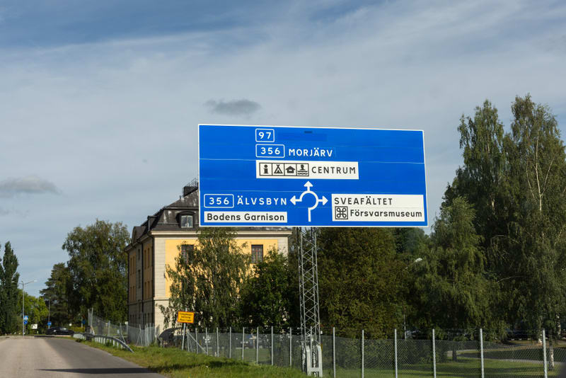 En stor blå vägskylt med vit text står i förgrunden och pekar mot olika destinationer som Morjärv, Centrum, Älvsbyn, Bodens Garnison, Sveafältet och Försvarsmuseum. Bakom skylten syns en gul byggnad med mörkt tak, träd, en grå himmel med moln och en