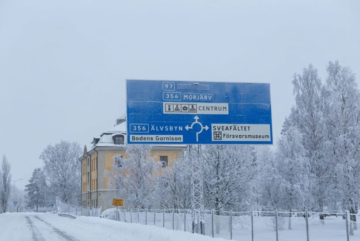 En vinterbild med en blå vägskylt i förgrunden, som visar vägbeskrivningar till olika platser. Snötäckta träd kantar vägen och en gul byggnad syns i bakgrunden. Marken är täckt av snö.