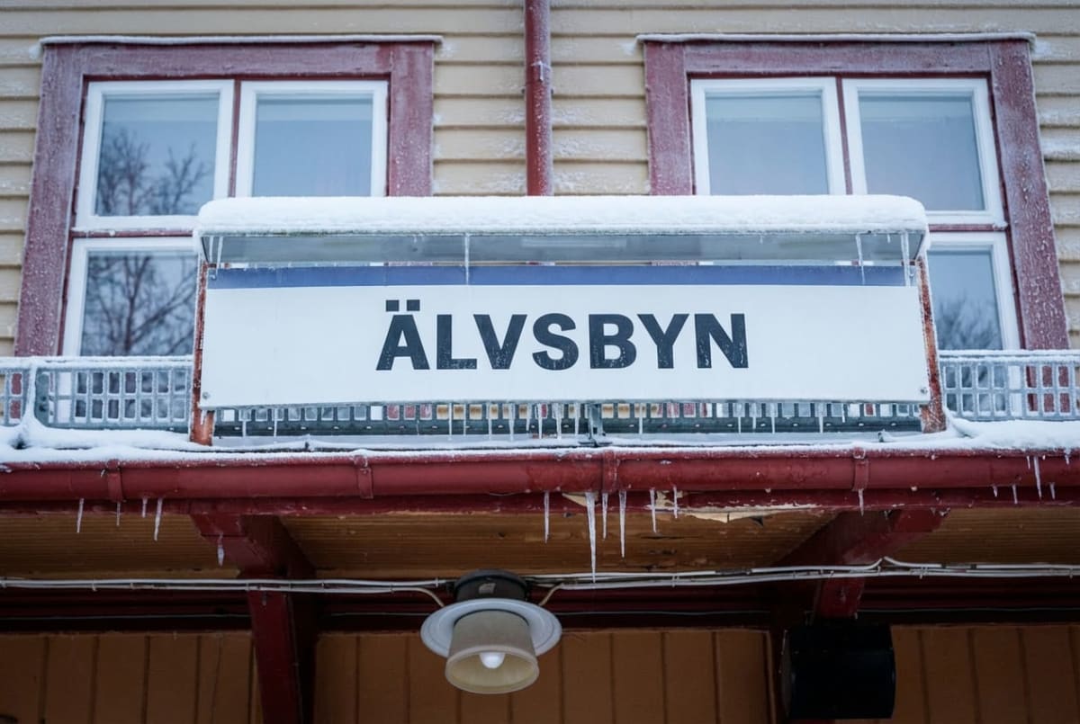 En fasad på en byggnad med en skylt som visar texten "ÄLVSBYN". Fasaden har liggande träpaneler målade i en ljus färg och röda fönsterramar. Det ligger snö på taket över skylten och det hänger istappar från takrännan. En lampa syns under takutsprånge