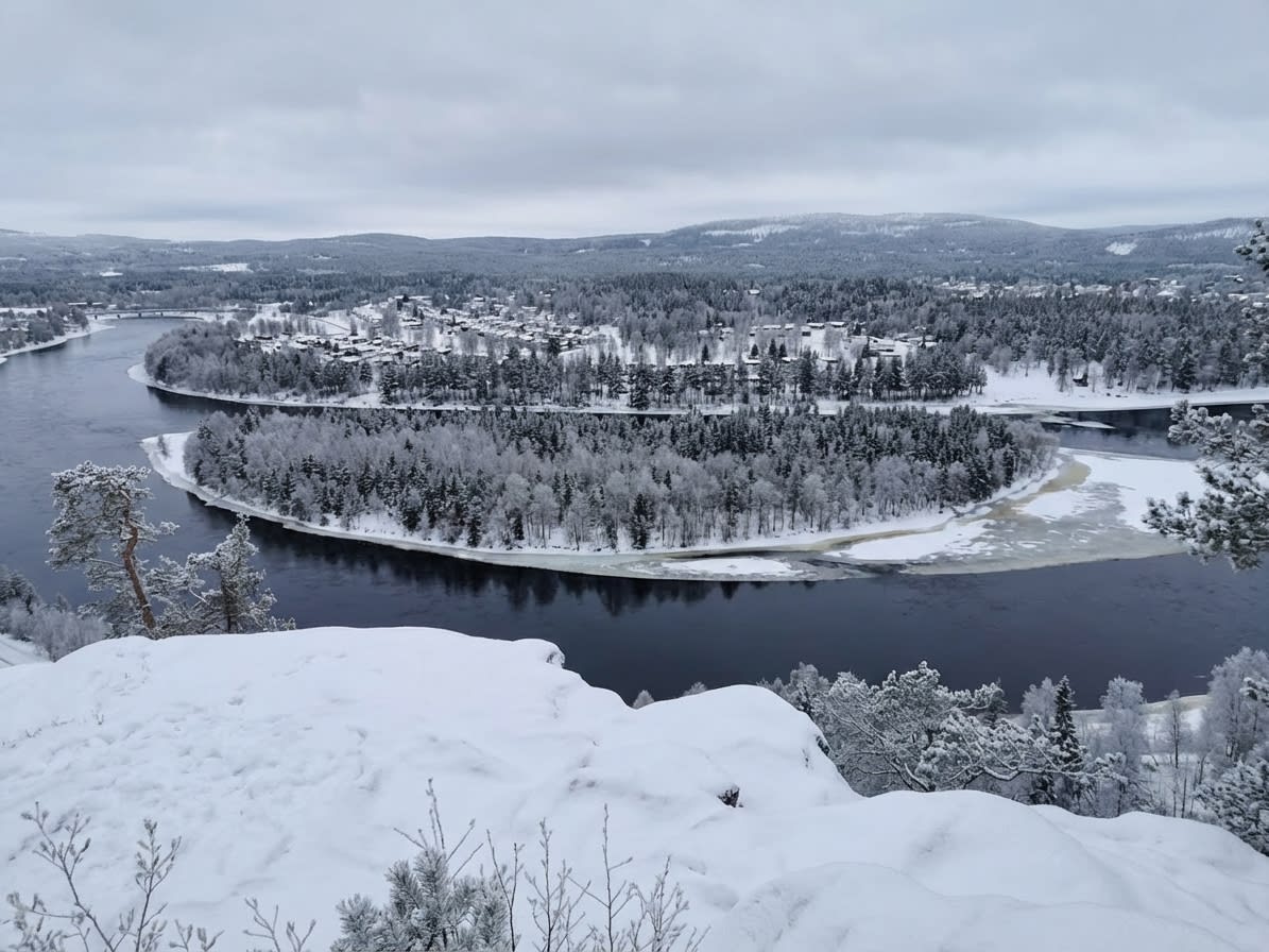 En vidvinkelbild tagen från en snötäckt höjd som visar ett vinterlandskap med en flod som slingrar sig genom snötäckta skogar och en bebyggelse. Delar av floden är isbelagda, och himlen är molnig.
