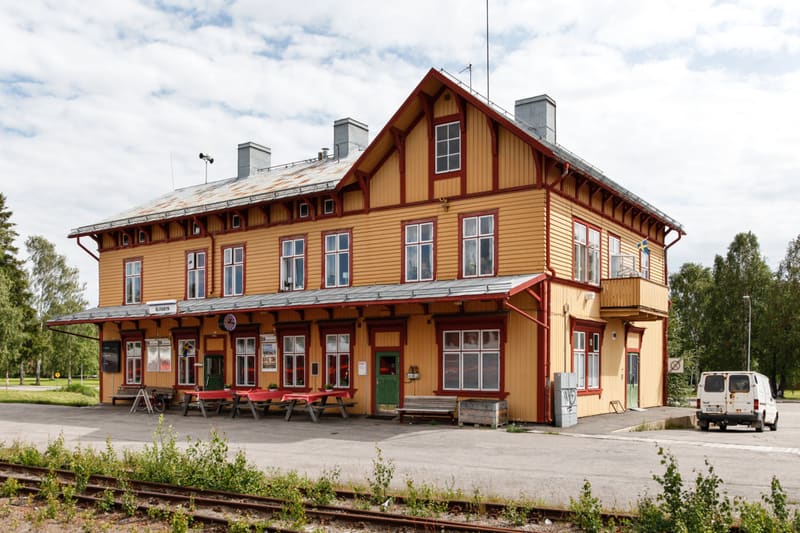 En gul trähusbyggnad med röda detaljer och vitbetsade fönsterkarmar står bredvid ett järnvägsspår under en molnig himmel. Byggnaden har en balkong med en svensk flagga, och utanför finns utomhusmöbler. En vit skåpbil är parkerad till höger om byggnad