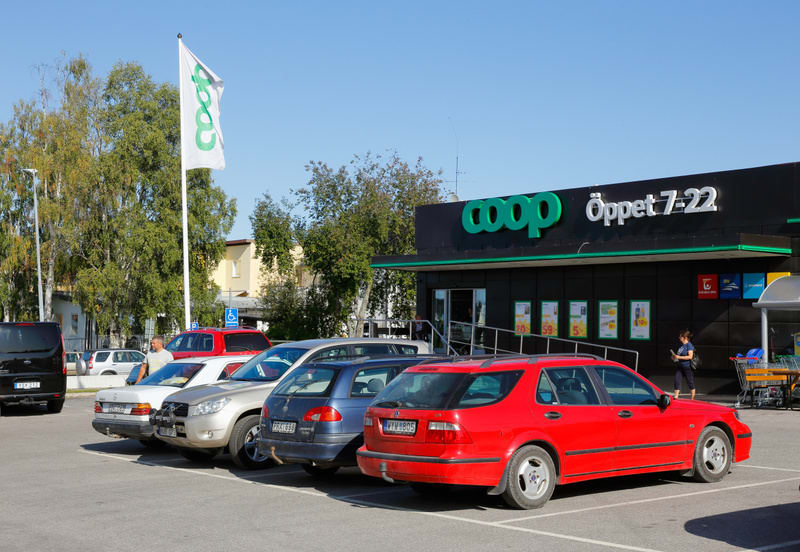 En parkeringsplats framför en Coop-butik under dagtid. Flera personbilar är parkerade. Till vänster syns ett högt flaggstång med en vit flagga med Coop-logotypen på. Butikens fasad är svart med grön text som anger "Coop" och "Öppet 7-22".