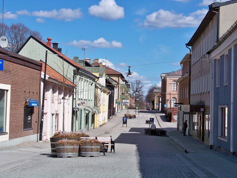 En gata med stenbeläggning i en småstad, kantad av flervåningshus med blandade fasadfärger. Husen har tak i rött och grått. Flera skyltar, möbler och gatlampor ses längs gatan. En person står vid en affärsingång och en fågel syns på trottoaren. En kl