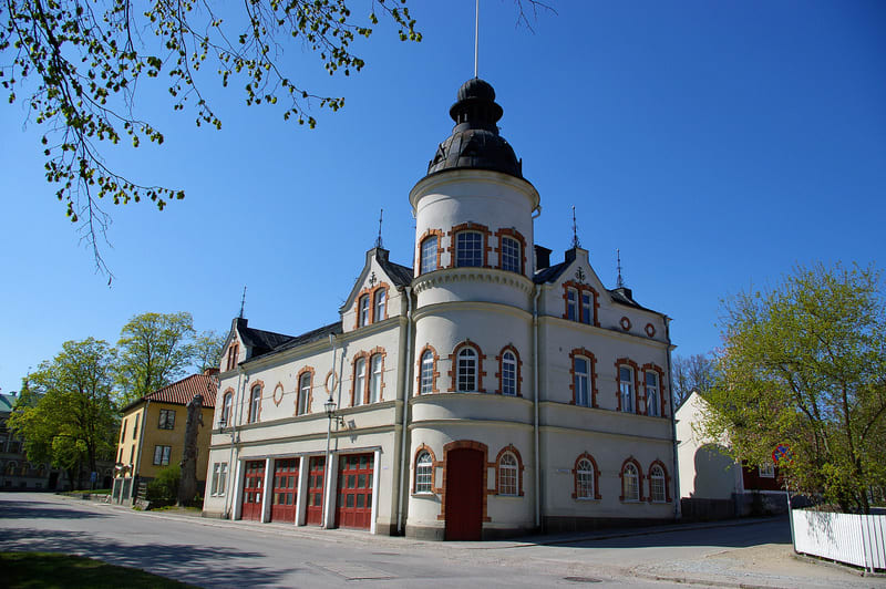 En stor, ljus byggnad med rött tak och en tornliknande del med rundade fönster. Byggnaden har garageportar på ena sidan och är omgiven av grönska och en väg. Himmelen är klarblå och några träd med nya löv syns.