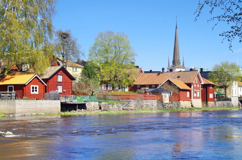En bild som visar en flod med rött vatten mot strandkanten och en rad röda trähus med fönster och tak, några med orangefärgade tak. I bakgrunden syns träd med gröna löv och en hög kyrkspira med klockor mot en klarblå himmel. Ett fågelbo är synligt i
