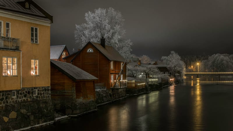 En vinterbild tagen på natten som visar en flodkant kantad av hus. Träden är täckta av snö eller frost, och ljus från fönster, utebelysning och gatlyktor reflekteras i vattnet. En bro med belysning syns i bakgrunden.