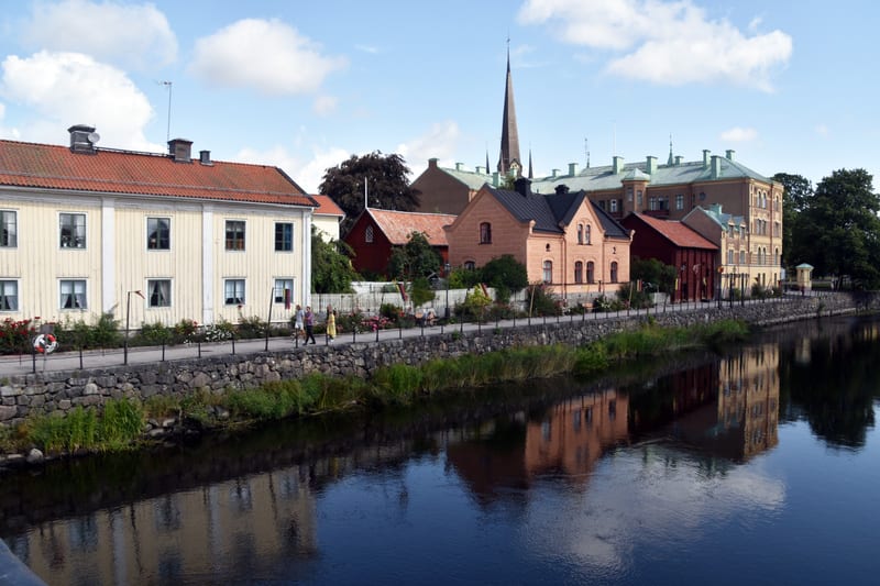 En bild som visar en flod eller å med bebyggelse längs ena sidan under dagtid. Flera byggnader i olika färger som gul, röd och sandfärg. Vattnet reflekterar byggnaderna och himlen, där moln syns. En kyrkspira sticker upp i bakgrunden. En gångväg med