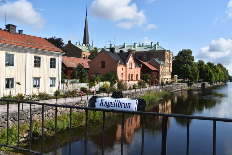 En bild tagen från en bro av en flod eller kanal som kallas Kapellbron. Längs vänster sida av vattnet syns äldre byggnader i olika färger samt grön vegetation och en stenmur. Ett kyrktorn syns i bakgrunden. Träd kantar vattnet på den högra sidan.