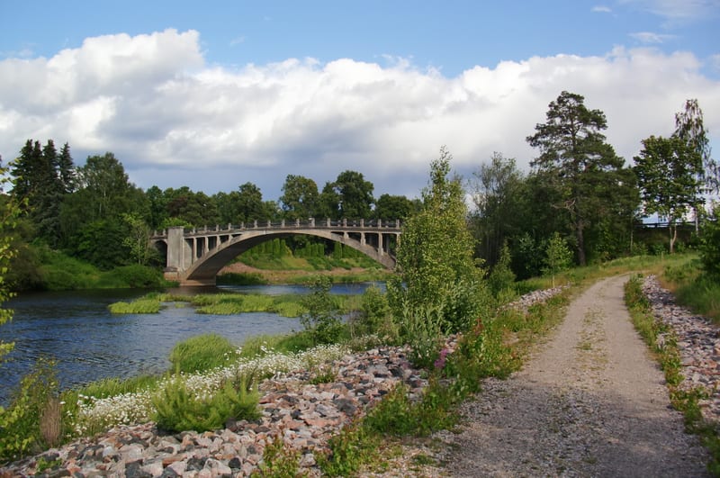 En betongbro sträcker sig över en flod omgiven av grönska med träd och buskar. En grusväg löper längs flodbanken på höger sida. Det är en ljus dag med moln på himlen.