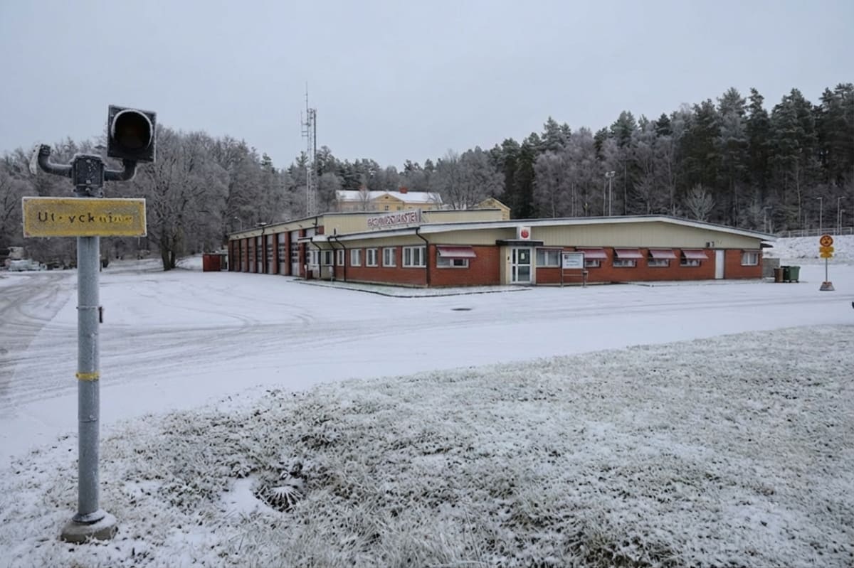 En byggnad med tegelväggar och ljusgul fasad, omgiven av snö. Ett varningsljus på en stolpe med texten "Ut-ryckning" står i förgrunden till vänster. Framför byggnaden finns en snötäckt yta och i bakgrunden skymtar en skog med snötäckta träd.