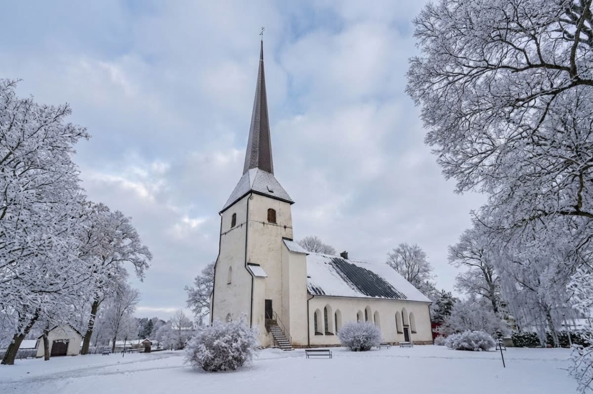En kyrkobyggnad med spetsig tornspira syns i mitten av bilden. Byggnaden är vitmålad med mörkt tak. Marken och träden runt omkring är täckta av snö, vilket indikerar vinterväder. Himlen är delvis molnig.