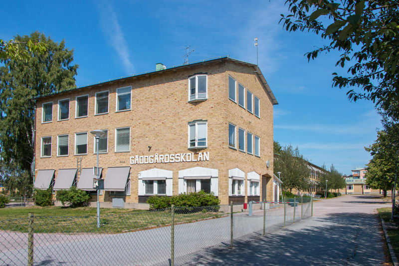 En gul tegelbyggnad med vita fönsterkarmar och texten "GÅDDGÅRDSSKOLAN" på fasaden, under en klarblå himmel. Framför byggnaden syns ett staket, gräs och buskar. Bilder visar även en gångväg och träd till höger.