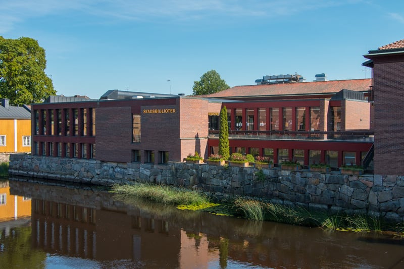 En modern byggnad med tegelväggar och stora fönster vid ett vattendrag. På fasaden står det "STADSBIBLIOTEK". Ett gult trähus med vita fönster syns till vänster. Blå himmel med några moln framträder i bakgrunden.