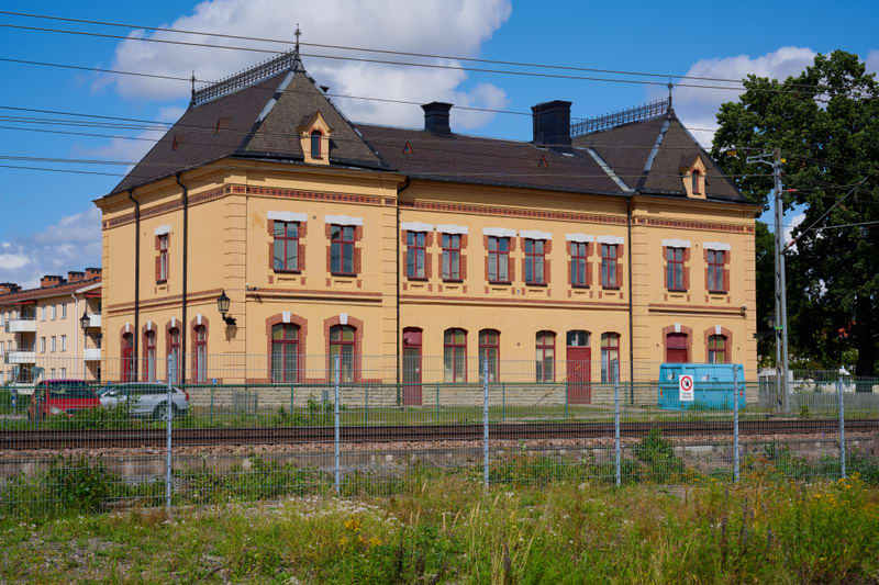 En gul byggnad med brunt tak och röda fönsterkarmar står bredvid ett järnvägsspår. I förgrunden syns ett gallerstängsel och gräs. I bakgrunden finns fler byggnader, träd och ett par bilar. Blå himmel med vita moln syns ovanför.