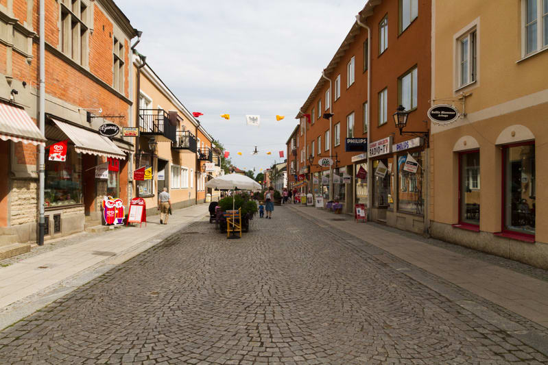 En gata med gatsten i en stadskärna, flankerad av byggnader med butiker på bottenvåningen. Flera personer promenerar på gatan och trottoarerna, och det finns uteserveringar med parasoller. Flaggor hänger tvärs över gatan, och himlen är molnig.