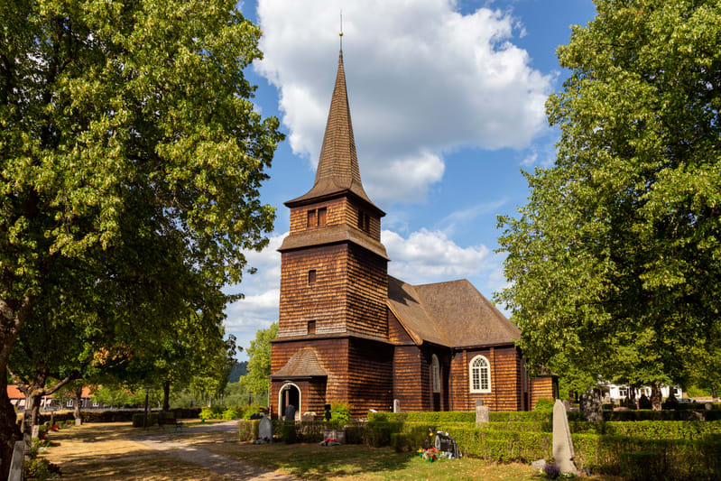 En rödbrun träkyrka med spetsigt torn, omgiven av stora gröna lövträd och en kyrkogård med gravar och tuktade häckar. En klarblå himmel med vita moln syns ovanför kyrkan och träden.