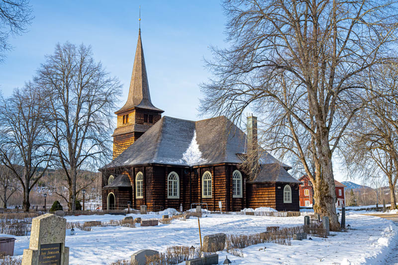 En stor träkyrka med en spetsig spira står i mitten av bilden. Marken är täckt av snö, och det finns gravstenar bland snön runt kyrkan. Nakna träd med kala grenar står runt kyrkobyggnaden. I bakgrunden syns röda byggnader och snötäckta kullar.