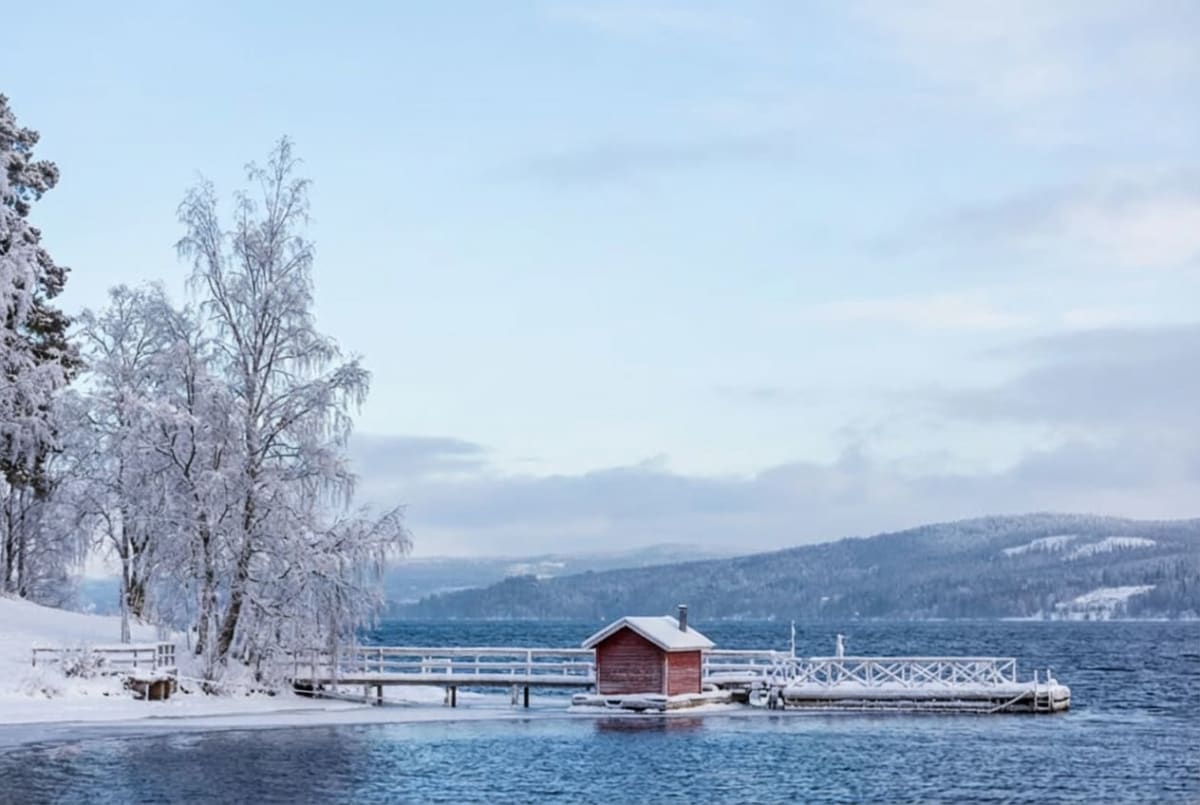 En vinterbild visar en sjö med öppet vatten och snötäckta stränder. En röd stuga står på en brygga vid vattnet, omgiven av snö. Ett par snöklädda träd står på land till vänster, och i bakgrunden syns snötäckta berg eller kullar under en ljusblå himme