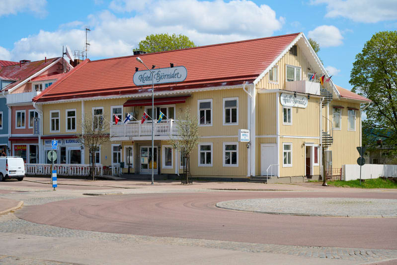 En gul tvåvåningsbyggnad med rött tak, skylten "Hotell Björnidet" syns på fasaden. På en balkong hänger flaggor från bland annat Sverige, Norge, Finland och Tyskland. Framför byggnaden finns en väg med en central refug och en skylt med bokstaven "P".