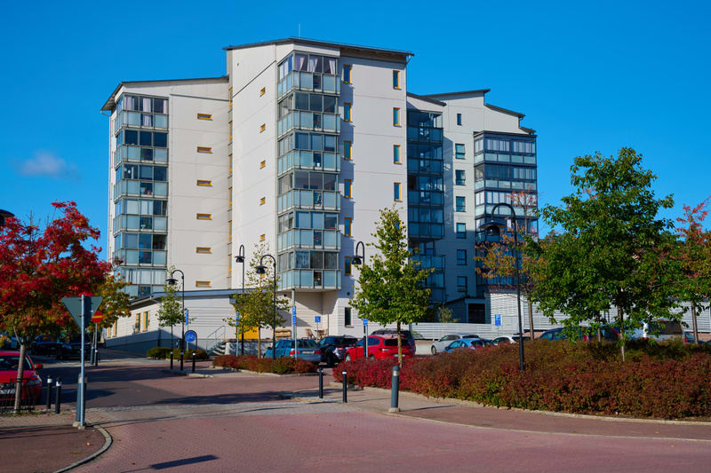 En bild utomhus som visar en modern, hög bostadsbyggnad i vit och grått med många glasinklädda balkonger under en klarblå himmel. Framför byggnaden finns ett grönområde med träd och buskar som delvis har höstfärger, samt en asfalterad väg med parkeri