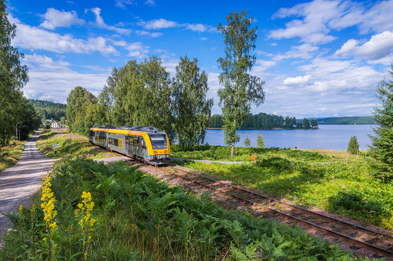 En gul och grå tågvagn står på en järnvägsskena vid sidan av en sjö, omgiven av grönska och träd. En grusväg löper parallellt med spåret. Blå himmel med spridda moln syns ovanför.