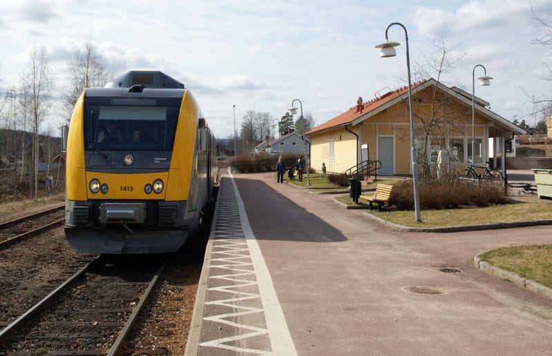 En gul och grå tågvagn står på ett dubbelspår vid en perrong. En gul byggnad med rött tak, en parkbänk och två gatlyktor syns bredvid perrongen. Flera personer står på perrongen och framför byggnaden. Träd och buskar med sparsam grönska omger området