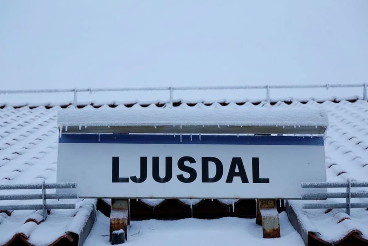 En vertikal bild visar en vit skylt med svarta versaler där det står "LJUSDAL", placerad framför ett snötäckt tak med takpannor och ett snöräcke. Snö och istappar syns tydligt på både skylten och taket, mot en ljus och molnig himmel.