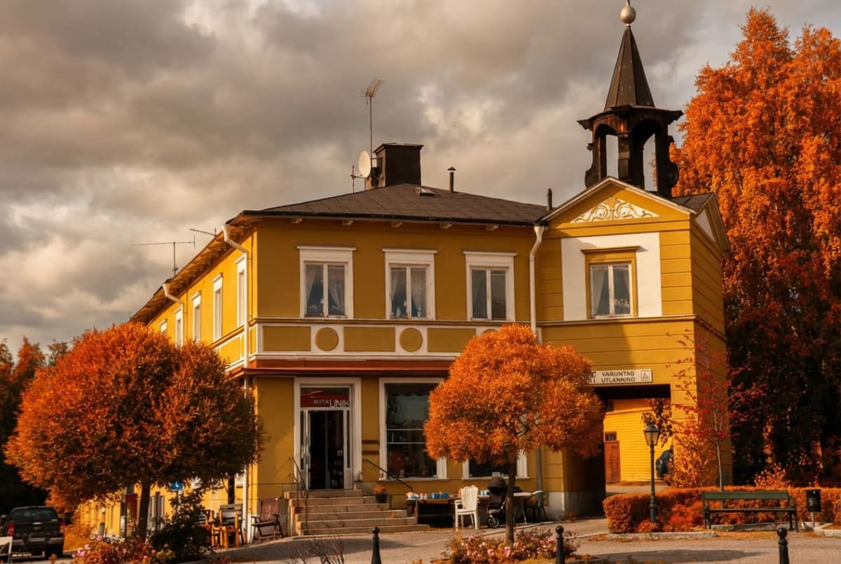 En gul byggnad med vita och bruna detaljer under en molnig himmel. Framför byggnaden står träd med orangea löv. En skylt med texten "VARUNTÅG UTLÅNNING" syns på byggnaden. På taket syns antenner och en spira.