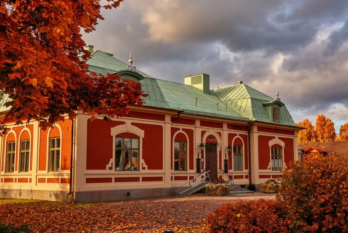 En röd tegelbyggnad med grönt tak och ljusa fönsteromfattningar under en molnig himmel. Framför byggnaden ligger nedfallna höstlöv, och träd med orangea löv syns i förgrunden och bakgrunden, vilket indikerar att det är höst.