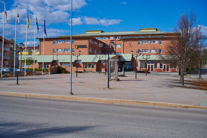 En fasadbild på en flervåningsbyggnad i tegel med grönmålade takdelar. Framför byggnaden finns en öppen plats med gatlyktor, flaggstänger med fyra flaggor och bar mark. Himmelen är blå med vita moln och ett par kala träd syns i bildens högerkant.
