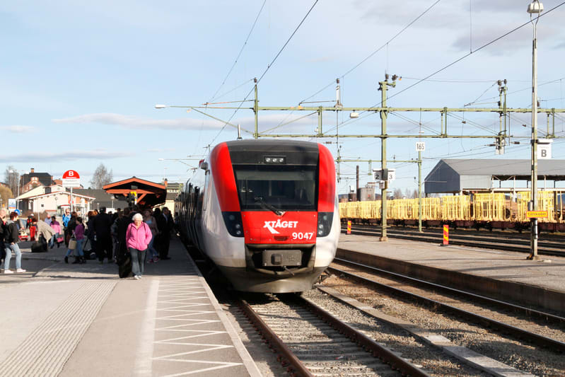 En röd och vit X-tågsmodell 9047 står på ett järnvägsspår vid en perrong där människor väntar och går. I bakgrunden syns bangård med godsvagnar med gula burar, samt en byggnad och träd. Blå himmel med tunna moln syns ovanför.