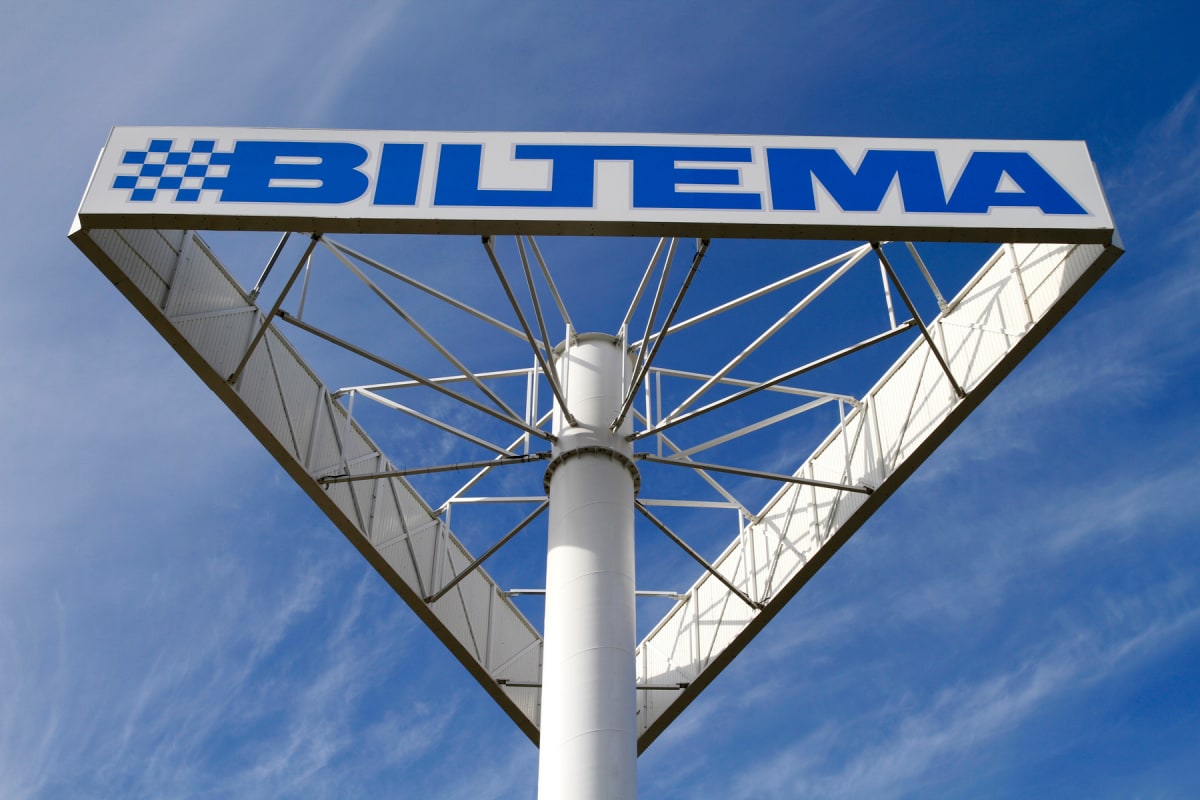 En stor skylt med texten "BILTEMA" i blått på vit bakgrund, mot en klarblå himmel med tunna vita moln. Skylten är fäst på en vit, rund stolpe med metallstöttor som strålar ut i ett triangulärt mönster bakom skyltytan. Bilden är tagen underifrån, vilk