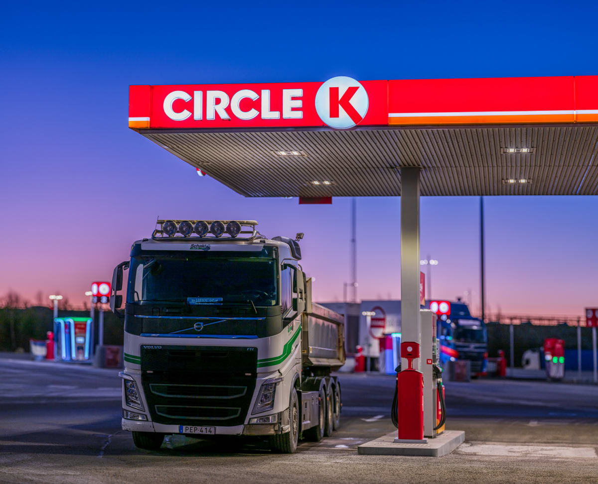 En stor vit lastbil med släp står parkerad under ett tak vid en Circle K bensinstation med en synlig Circle K-skylt. En bensinpump syns intill lastbilen. Bakgrunden har en blåviolett himmel med ett fåtal ljus.