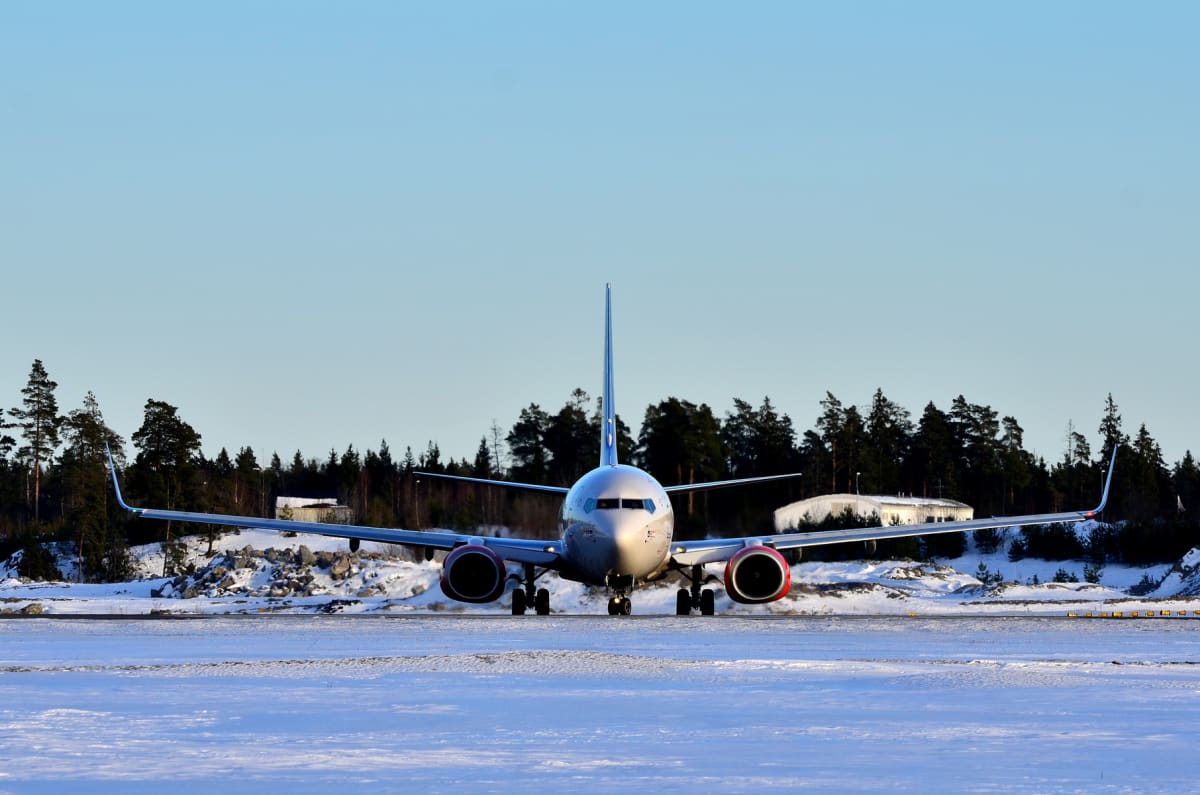 Ett flygplan med rödmarkerade jetmotorer står på en snötäckt landningsbana under en klarblå himmel. I bakgrunden syns skog med barrträd och snöklädda klippor, samt en vit byggnad till höger om flygplanet.