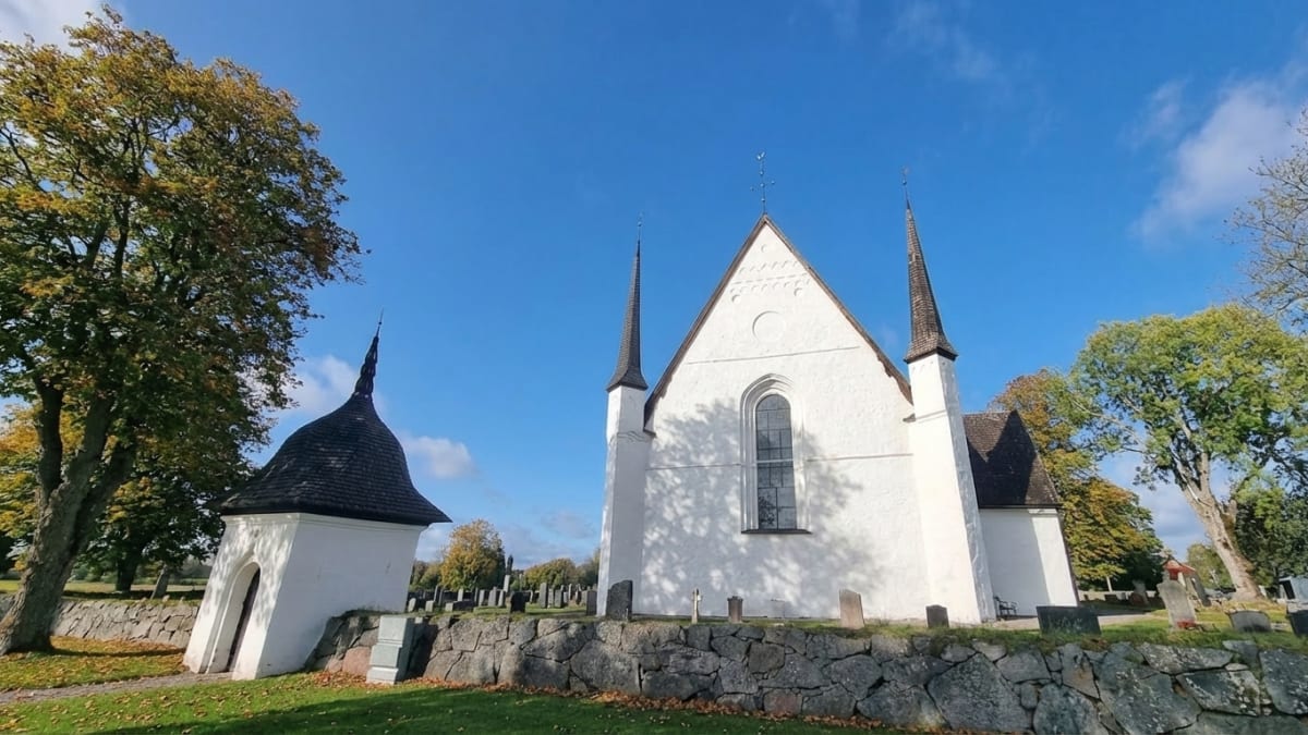 Bild på tierp kyrka med ett större centralt torn och två mindre torn på vardera sidan. Till vänster om kyrkan syns en vit byggnad med ett klockformat mörkbrunt tak. Kyrkan omges av en stenmur och ett rektangulärt grönt gräsområde. Flera träd med gulg