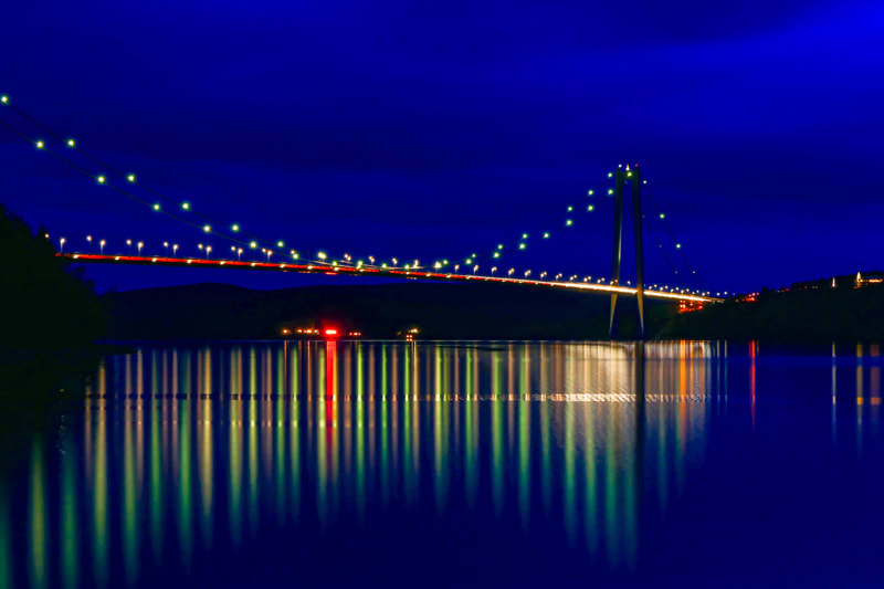 En bro fotograferad i mörker, med ljusbelysta kablar som reflekteras i vattnet undertill. Vänster sida av bron har grönskande träd i förgrunden, medan höger sida har ljus från byggnader i bakgrunden. Ett trafikflöde är synligt på bron.