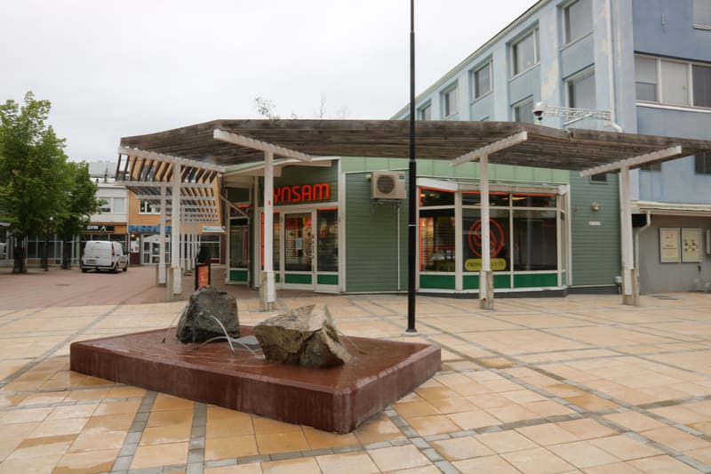 En bild av ett torg med en fontän i förgrunden, en butik med skylten "SYN SAM" och vitpersienner i bakgrunden. Ett vitt servicefordon står till vänster, och byggnader med fönster och en luftkonditionering är synliga.