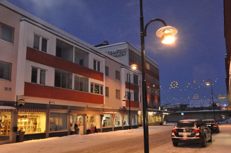 En gatsyn i Kramfors under kvällstid eller tidig morgon. Gatan är täckt av snö och det syns byggnader med butiker på bottenplan. En skylt på en byggnad lyder "KRAMFORS KOMMUN". Gatubelysning och dekorationsbelysning lyser upp scenen.